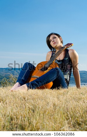 Beautiful female guitarist sitting and posing with acoustic guitar on field against blue sky.