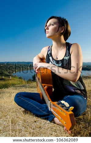Beautiful female guitarist sitting and posing with acoustic guitar on field against blue sky.