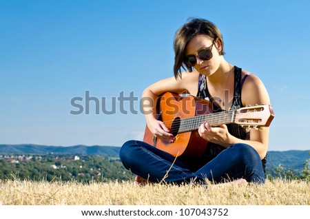 Beautiful female guitarist playing acoustic guitar on field against blue sky.