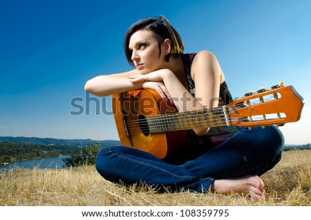Beautiful female guitarist sitting and posing with acoustic guitar on field against blue sky. Wide angle.