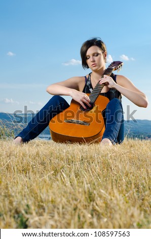 Beautiful female guitarist playing acoustic guitar on field against blue sky