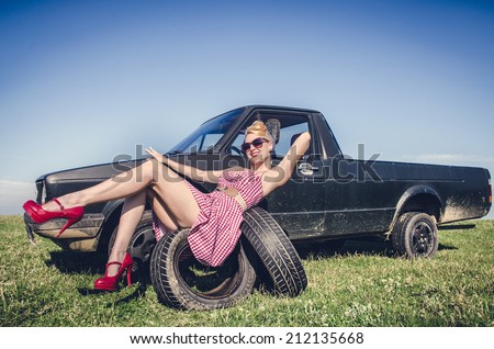 Sexy pin-up girl in short dress sitting on tires next to retro pickup car