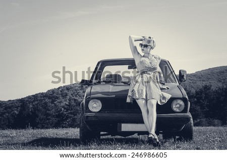 Female model in short dress posing next to retro car on a field. Grayscale image.