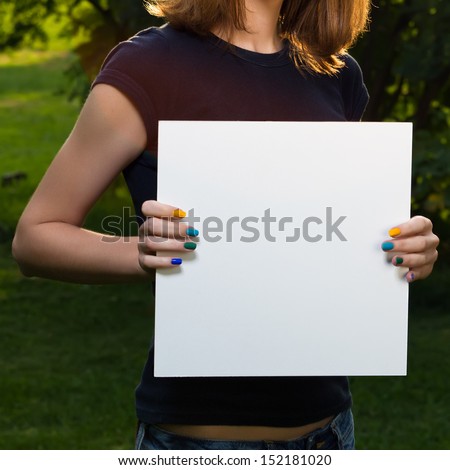 Young girl holding white blank paper against background of summer green park. Young girl showing blank board.