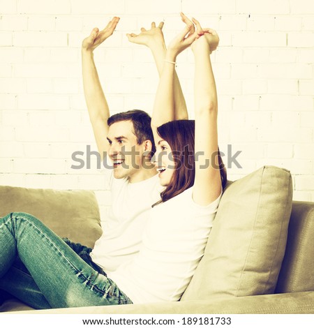Happy couple with arms raised up. Young couple watching TV on sofa. Toned image