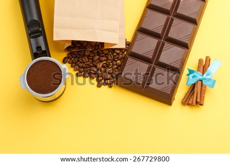 Overhead view of coffee machine filter holder, coffee beans, bar of chocolate, cinnamon on yellow background