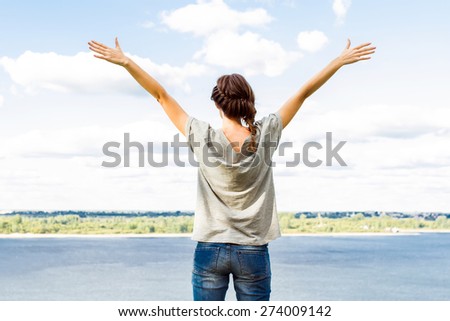 Rear shot of young woman raising arms up towards blue sky and river.