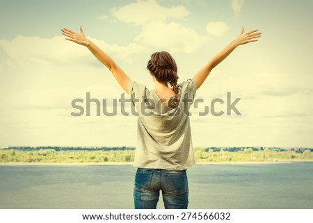 Rear view of young woman raising arms up towards blue sky and river.