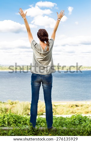 Rear full length view of young woman raising arms up towards blue sky with clouds and river.