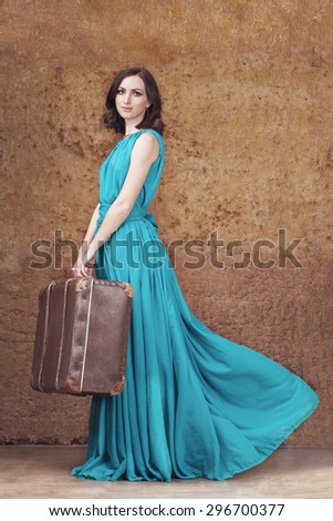 Full length portrait of young woman in elegant blue dress holding vintage suitcase. Travel concept