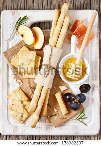 Grissini bread sticks, crackers with salt, rosemary honey, prosciutto ham, apple and grape on a white cooking board.