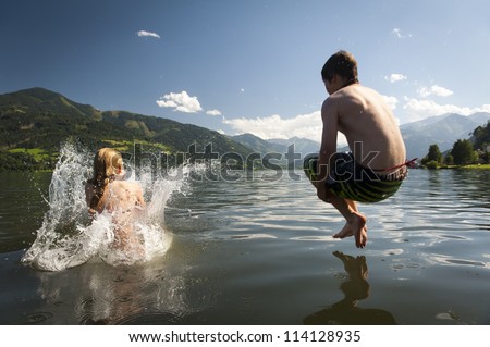 girl already in the splashing water and boy in the air while they where jumoing into a lake, with nice nature and mountains in the back