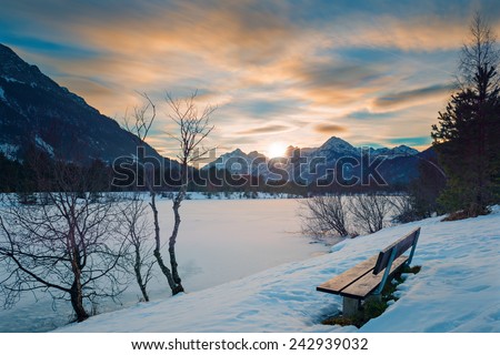 lonely 	park bench at sunset in alps with frozen lake in winter