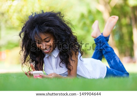 Smiling teenage black girl using a phone, lying down on the grass - African people