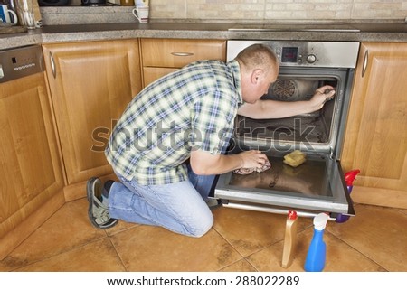 Man kneels on the floor in the kitchen and cleans the oven. Cleaning work in the home. Man helping his wife with maid service.