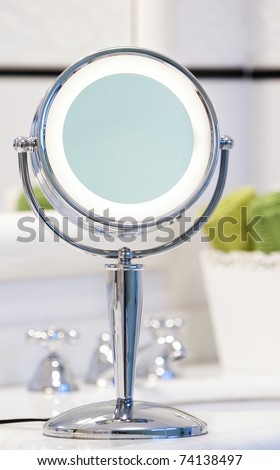 Round chrome lighted makeup mirror in bathroom. Green towels in the background.