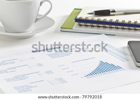 Sheet of paper with figures, tables and graph, small notebook and pen, cell phone and a cup of tea