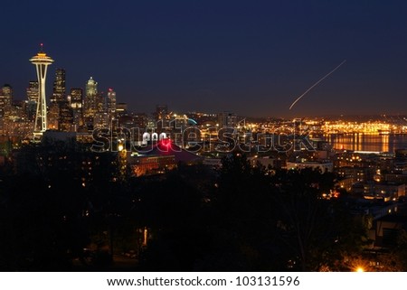 Downtown Seattle at twilight. The Space Needle on the left of frame with Elliott Bay on the right. The streak of light in the sky is a plane landing at the airport. Washington State, USA.