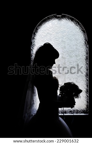 Silhouette of bride besides the window with her wedding bouquet.