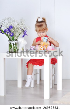 Little girl in red dress sits at table and waters of the cup doll.