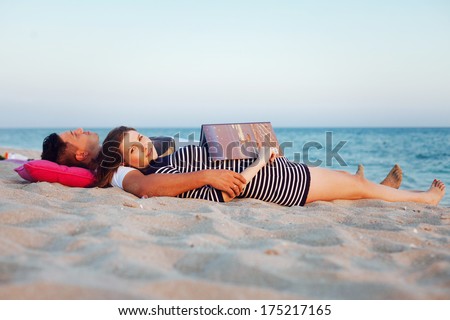 Young beautiful couple have a picnic on the beach. Beautiful pregnant woman reading a book of fairy tales.
