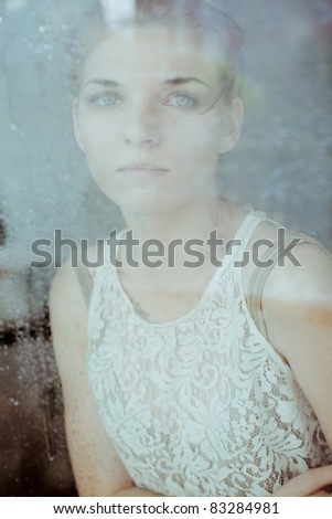 Beautiful sad girl standing at the window watching the rain