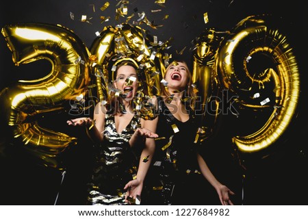 Beautiful Women Celebrating New Year. Happy Gorgeous Girls In Stylish Sexy Party Dresses Holding Gold 2019 Balloons, Having Fun At New Year\'s Eve Party. Holiday Celebration