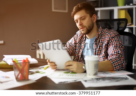 Thoughtful male person looking to the digital tablet screen while sitting in modern loft interior at the table, experienced entrepreneur reading some text or electronic book at the office