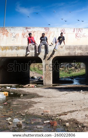 CAPE TOWN, SOUTH AFRICA - JANUARY 31: Three young township men sit and talk on a bridge with filthy water running underneath in a township, in Cape Town, South Africa, on January 31, 2012