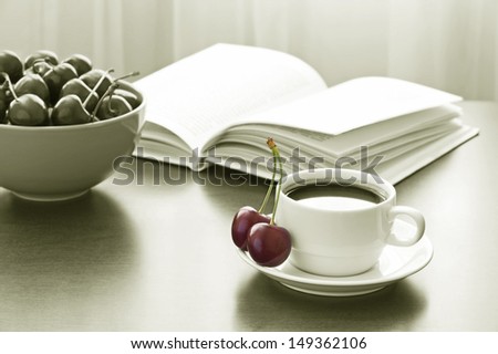 Cup of coffee, book and bowl of sweet cherries on table against window. Black-and-white