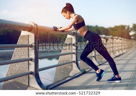 Attractive and strong woman stretching before fitness and listening to music with headphones on the lake in the summer. Sports concept. Healthy lifestyle