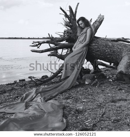 portrait image of sad model girl wrapped in fabric standing near fallen tree with head down on the beach
