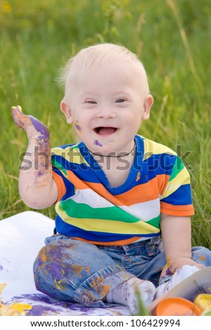 Little baby boy with Down syndrome painting finger paints on white paper with a smile. Happiness is in creativity.