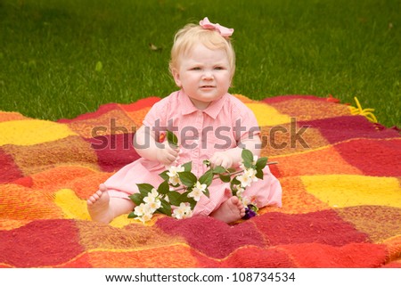 Baby girl wearing a pink dress and a bow in her hair sitting on a plaid on the lawn with flowering jasmine twig in the hands