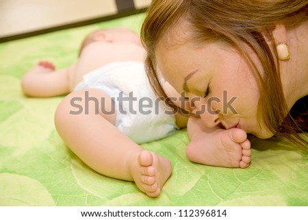 Pretty young mother kissing the tiny feet of her newborn baby who is sleeping in bed