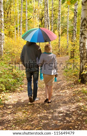 Happy pregnant woman and a man walking in the autumn forest under a rainbow colorful umbrella, rear view