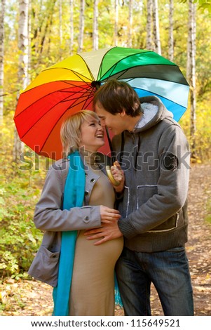Happy pregnant woman and a man walking in the autumn forest under a rainbow colorful umbrella and kissing
