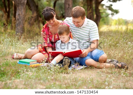 Happy father, mother and son reading a book on the lawn in the park. Family reading together.