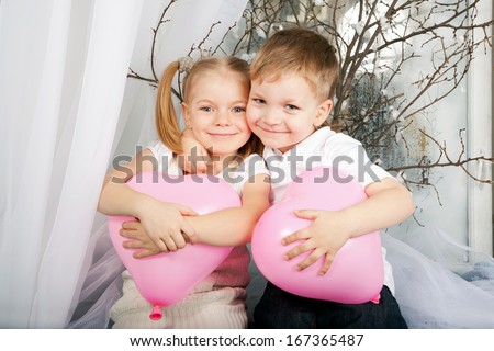 Little boy and girl romantic couple in love. Little kids hugging and holding heart balloons, sitting on the window sill. Valentine\'s Day and love concept, ready for your logo, text or symbols.