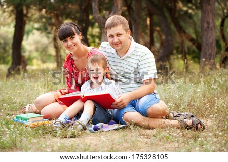 Happy Father, Mother And Son Reading Book On The Grass In The Park. Family Reading Together.