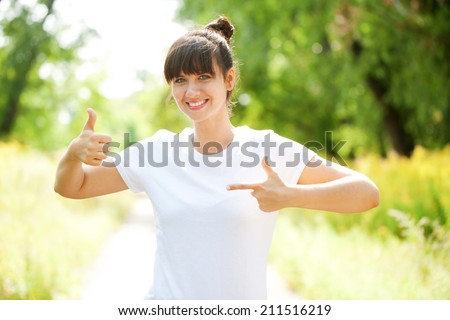 Woman in white t-shirt pointing at something and showing a thumbs up sign outdoors. Ready for your logo, text or symbols.