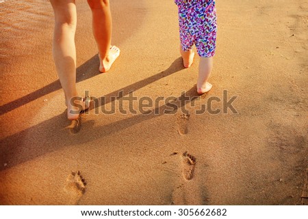 Mother and baby walking along the beach. Legs, feet and footprints on the sand. Summer vacation.