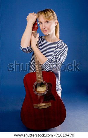 Beautiful middle aged blonde woman in a striped vest posing with a red guitar. Isolated on blue.