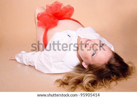 Beautiful pregnant woman with blond hair and blue eyes smiling. In the pregnant tummy red bow. Isolated on a beige background.