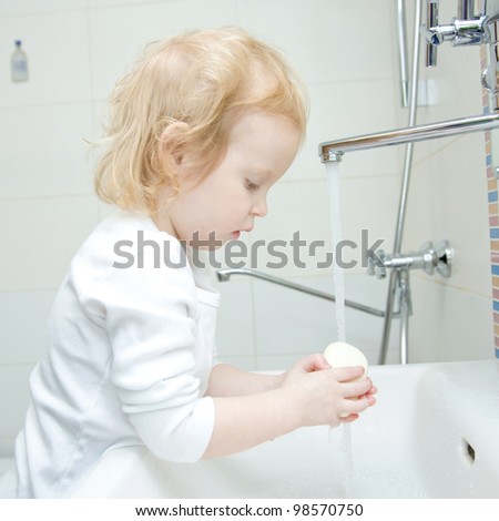 The little blonde smiling girl washing hands and face with soap in the bathroom. Hygiene. The girl wearing a blank white shirt. Ready for your design or logo
