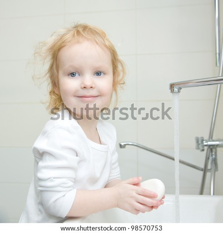 The little blonde smiling girl washing hands and face with soap in the bathroom. Hygiene. The girl wearing a blank white shirt. Ready for your design or logo
