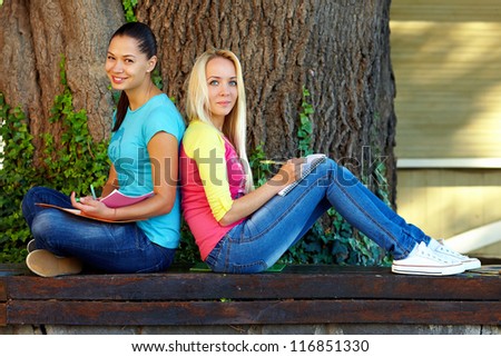 two smiling female student sit on bench under an old tree