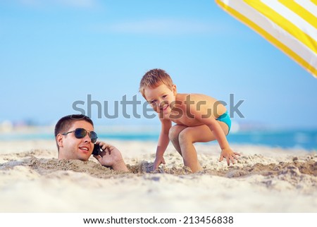 kid playing around father\'s head in sand, talking on the cell phone