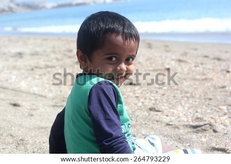 smart young happy indian boy with big eyes sitting at a beach turning back and smiling on a bright beautiful sunny day with calm blue waves in the background
