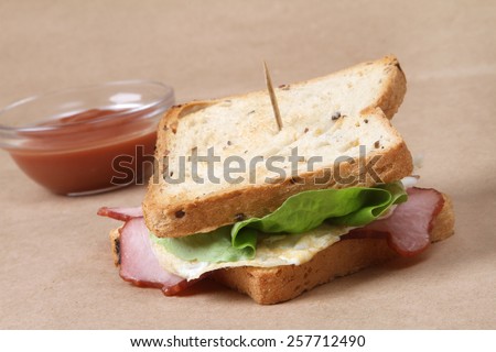 Club sandwich with ham and pork fried bacon, baked egg topped with cheese and lettuce next to a glass bowl of fresh hot red tomatoes ketchup